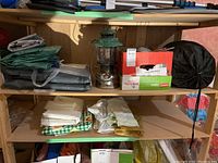 Two wooden shelves holding camping equipment including tarps, tablecloths, sleeping bag, Coleman lantern, and a box.