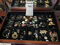 Open velvet-lined jewelry box showing multiple compartments filled with earrings, brooches, pendant, and watch