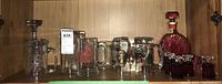 Shelf with glass decanter with deep red cranberry coloration and stopper, several beer mugs including some with logos or painted designs, small cranberry to clear shot glasses grouped together.