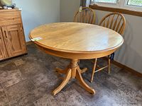 Round wooden pedestal dining table shown in a room with floor tiles and two wooden chairs against the wall.