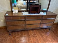 Full front view of the wood dresser showing all nine drawers and attached mirror, floor and wall visible.