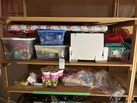 Two wooden shelves holding assorted gift wrapping supplies including multiple rolls of Santa-themed gift wrap, clear plastic totes filled with bows and ribbons, white boxes, and gift bags.