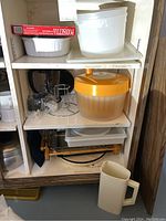 Three shelves showing various kitchen items including plastic pitchers, lettuce spinner, glass baking dish in wooden holder, CorningWare casserole dish