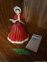 Front view of the Royal Doulton figurine in red dress holding a book, on wooden surface with maxsold tag and size indicator (6.5 inches).