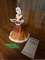 Full view of the Royal Doulton figurine standing 6.5 inches tall, dressed in a brown to burnt orange gradient dress with white trim, holding a book.