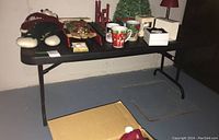 Table with black resin top and folding black metal legs shown with various Christmas decorations on it. Floor in basement setting.