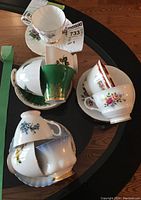 Nine assorted tea cups and saucers arranged on a round wooden table showing various floral patterns, one green gold accented cup, and delicate porcelain material.