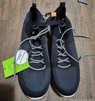 Top-down photo of the pair of men's black athletic shoes size 10 placed side by side on a textured surface. Shoes have mesh fabric upper, white and black laces, and pull tabs at heel and tongue. Green price tag attached indicating new condition.