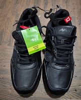 Pair of new black men's athletic shoes with laces and tags, size 12, front view on wooden floor.