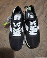 Pair of women's black and white athletic shoes, size 7, showing the top, sides, and laces intact, placed on wooden floor.