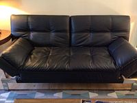 Black faux leather futon couch with tufted stitching, arm rests in upright position