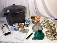 Photo shows black speckled enameled large canning pot with lid, metal jar lifter, multiple packs of flat jar lids and jar rings, green funnel, and metal mesh strainer.