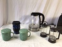 Lot arranged on a cloth backdrop, showing all items: electric kettle, two French presses, two green ceramic mugs with lids, and blue travel mug.
