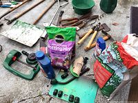 Overview of assorted gardening tools, potting mix bags and watering equipment arranged on concrete floor.