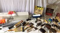 Wide view of assorted painting items including brushes, rollers, sanding blocks, plastic sheeting, and drop cloth packs arranged on a white surface with white background.