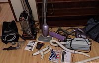 Full lot overview showing Shark vacuum, steam mop, handheld steamer, Oreck canister vacuum, attachments, and manuals on floor