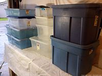 Photo showing a collection of plastic storage totes of various sizes stacked on a table, including gray, clear, and blue colors.