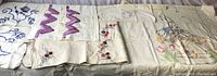 Photo showing a variety of linens with detailed needlework, lace, and embroidered floral and character designs spread out on a surface.
