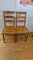 Front view of two Ikea pine dining chairs side by side on hardwood floor under a window, showing overall shape and condition.