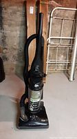 Front view of black Dirt Devil upright vacuum with clear dust canister and hose attached, placed on basement floor near metal bed frame and wooden shelf.