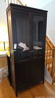 Front view of Ikea Hemnes display cabinet with glass doors and lower solid panels, black-brown finish, against a staircase background.