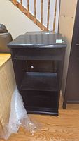 Front view of black laminate entertainment stand showing three shelves and wheels, bubble wrap on floor nearby.