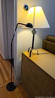 View showing both black metal goose-neck floor lamp with round base and black cast iron desk lamp with white cloth shade on a wooden cabinet near a couch and door.