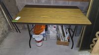 Full view of portable folding table with wood grain laminate top and black metal folding legs, placed in basement with some surrounding storage.