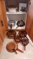 Cabinet shelf with vintage Corning Visions amber glass cookware including casseroles, frying pans, and rectangular lid with aluminum pot and white drip coffee maker below.