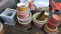 Photo showing multiple stacks of terracotta and ceramic pots, saucers, and planter bottoms on a wood floor outdoors.