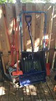 Various snow shovels and snow scoop propped against a wooden fence showing handles and scoop blades