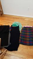 Three skirts laid flat on floor showing black, pleated black, and multi-color plaid skirts.