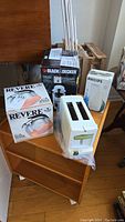 Photo showing boxed Revere copper kettle, Black & Decker coffee maker box, Philips steamer box, and white Philips toaster placed on wooden shelf.