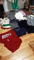 Overview of assorted folded sweaters, vests, and sweatpants including a red Roots Athletics sweatpant, knit sweaters in cream, navy, gray, black, and other colors piled on a wood floor.