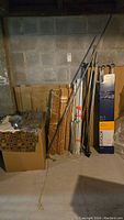 Wide shot showing several bamboo roll-up blinds, curtain rods, and some fabric panels stacked next to a cardboard box in a basement with concrete walls.