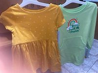 Two toddler dresses, one yellow with a heart pattern and one green with a rainbow graphic and front pouch pocket, hanging on hangers.