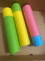 Three foam pool toys in green/yellow, yellow, and pink/blue colors shown from the side in a cardboard box.