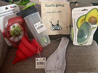 Photo showing all six kitchen gadgets together on a green surface, including apple corer and slicer, two collapsible funnels, shaker cup, garlic bag, spoon rest, and avocado container.