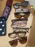Top-down view of 8 pairs of eyeglasses laid out in a cardboard box with two different new eyeglass cases visible at the side, one black with white polka dots and the other pink paisley.