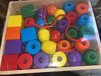 Full view of all wooden beads in a wooden tray with transparent cover, showing variety of shapes and colors and red lacing string.