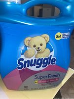 Blue plastic bottle of Snuggle SuperFresh fabric softener, showing front label with teddy bear logo and 5in1 odor elimination technology.