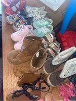 Top view of 11 pairs of toddler shoes in different styles & colors arranged on a wooden surface. Shows a variety of styles including red glitter flats, patterned sneakers, suede boots, and other shoes.