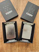 Photo showing two new in box Zippo brand lighters with chrome finish in their original black display boxes with Zippo logos.