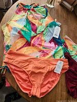 Photo showing a bright multicolor leaf patterned bikini top and two bikini bottoms (peach and wine color) with tags attached, laid flat on a wooden surface.