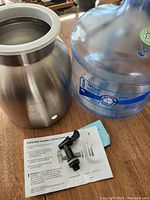 Water jug made of clear Tritan plastic next to stainless steel holder and assembly instructions with small dispenser faucet piece.