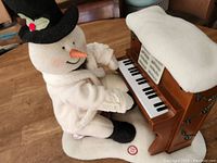 Soft fabric snowman figurine dressed in white, wearing black top hat with holly detail, playing piano with miniature sheet music and white cover on piano top.