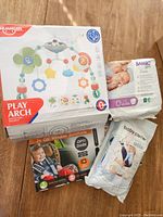 Photo showing boxed colorful baby play arch mobile, white packaged baby carrier, pack of size 2 Bambo Nature diapers, and box containing two dark car window sunscreens with suction cups