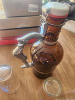 Amber glass growler with unique metal figure handle and clamp lid on wooden table with two small glasses.