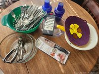 Photo shows a variety of kitchen items on a wooden table. Includes green plastic colander holding flatware, two clear and white glass bowls, pie pan with purple and yellow lid, handheld cheese grater, two blue shakers, and metal utensil.