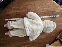 Porcelain doll lying on stomach on wooden table with a measuring tape, showing the back of the winter outfit jacket and pants.